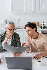 young man pointing with hand near thoughtful dad holding folder with documents while sitting at laptop