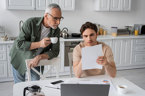 serious young man looking at document near laptop and senior dad with coffee cup