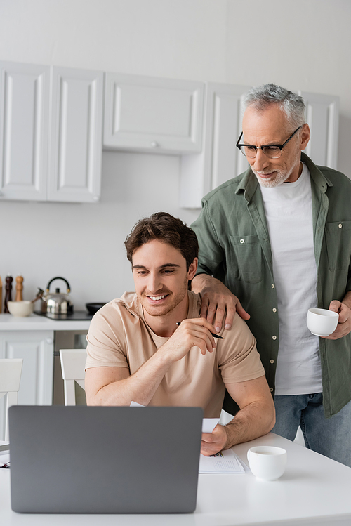 mature man with coffee cup touching shoulder of young son smiling near laptop in kitchen