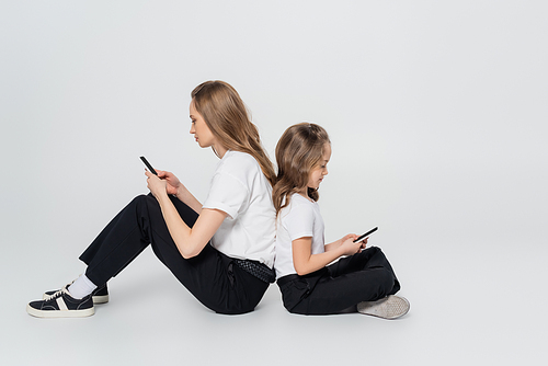 side view of mother and child using smartphones while sitting back to back on grey background