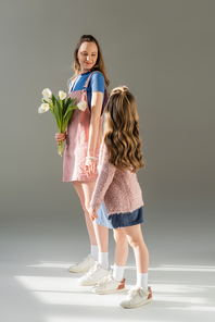 full length of happy mother holding flowers and looking at daughter on grey