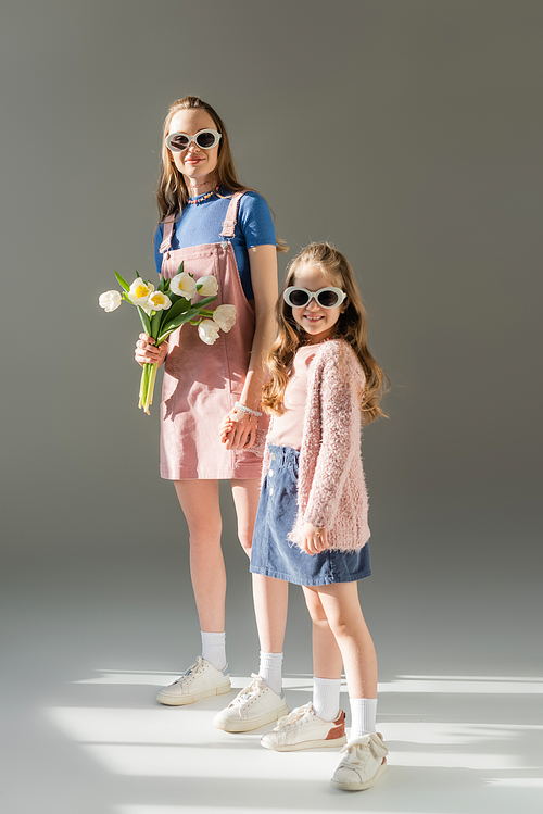 full length of happy mother with flowers and daughter in sunglasses holding hands on grey