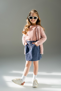 full length of happy kid in sunglasses standing with hands in pockets on grey