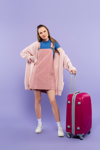 full length of woman in pink overall dress standing with hand on hip near luggage on purple