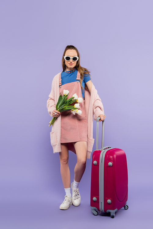 full length of woman in pink overall dress and sunglasses standing with flowers near luggage on purple