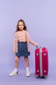 full length of cheerful girl standing with pink luggage on purple