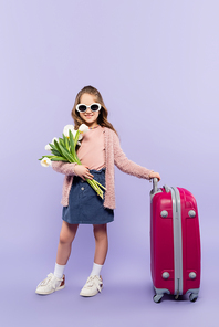 full length of cheerful girl in sunglasses standing with flowers near pink luggage on purple