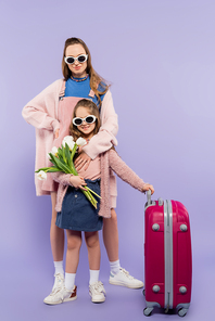 full length of mother and happy child in sunglasses standing near luggage on purple