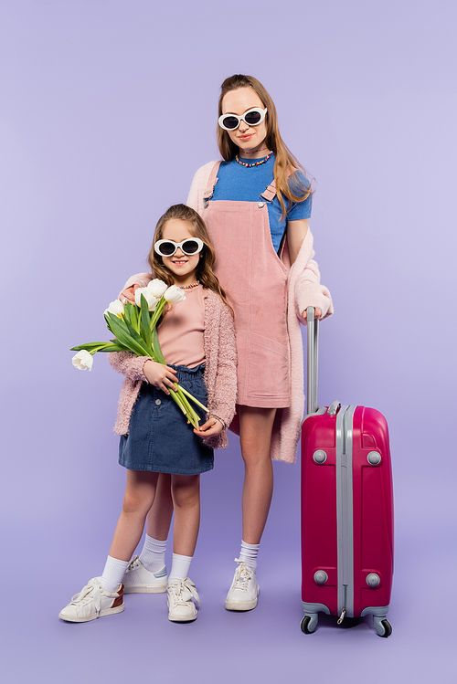 full length of mother and child in sunglasses holding flowers and standing near baggage on purple