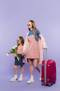full length of mother and child in sunglasses holding hands and walking with baggage on purple