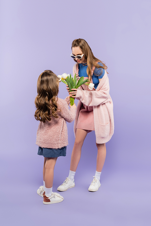 full length of child in sunglasses giving flowers to mother in overall dress on purple
