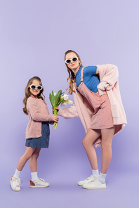 full length of happy girl in sunglasses giving flowers to mother in overall dress on purple