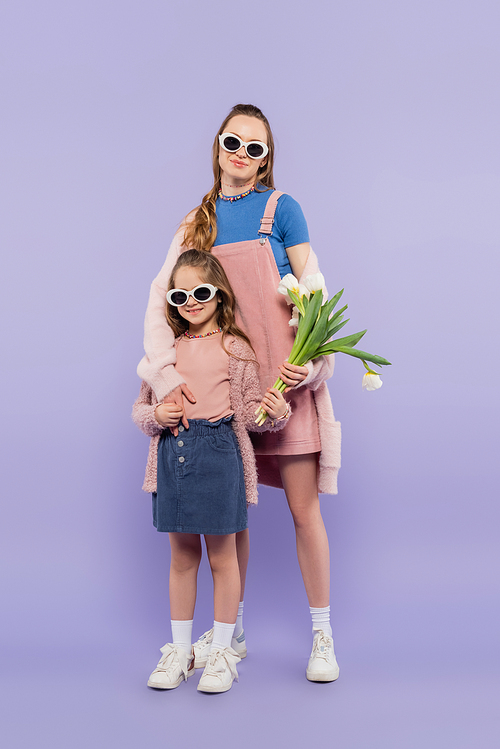 full length of child in sunglasses holding flowers near mother in overall dress on purple