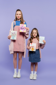 full length of mother and happy daughter holding greeting cards on purple, mothers day concept