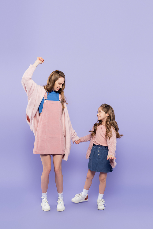 full length of happy mother in pink overall dress holding hands with joyful girl on purple