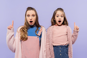 shocked mother and daughter pointing with fingers up and looking at camera isolated on purple