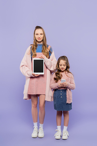 full length of woman showing digital tablet with blank screen near kid using smartphone on purple