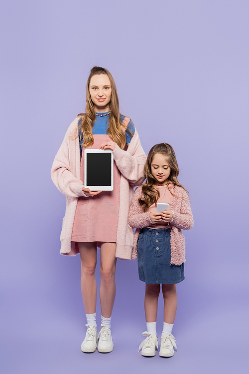 full length of woman showing digital tablet with blank screen near kid using smartphone on purple