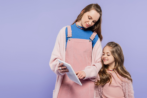 smiling woman showing digital tablet to happy daughter isolated on purple