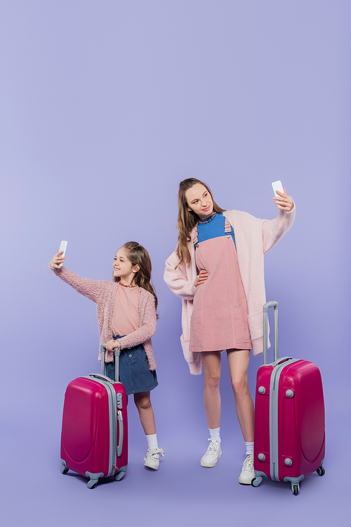 full length of happy mother and child taking selfie on mobile phones near baggage on purple