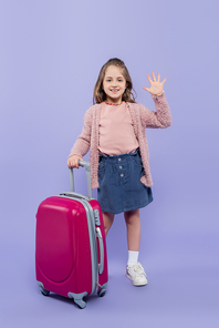 full length of happy girl waving hand near pink baggage on purple