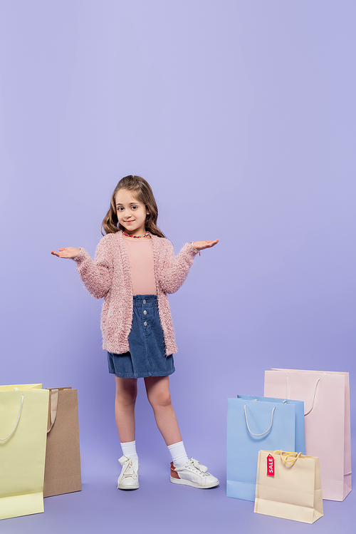 full length of happy kid pointing with hands near shopping bags on purple