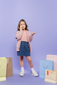 full length of happy kid talking on smartphone and standing near shopping bags on purple