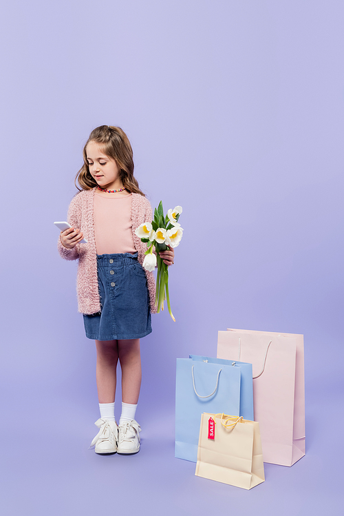 full length of kid holding flowers and using smartphone while standing near shopping bags on purple