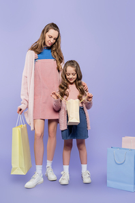 full length of kid looking inside of shopping bag near mother on purple