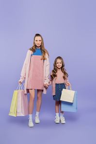full length of pleased mother and daughter holding shopping bags on purple