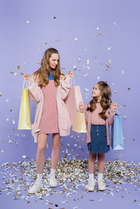 full length of mother and daughter holding shopping bags near falling confetti on purple