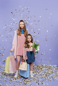 full length of happy mother and daughter holding shopping bags and flowers near falling confetti on purple