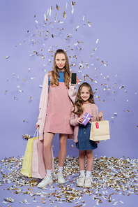 full length of happy mother holding smartphone and shopping bags near daughter with present near falling confetti on purple