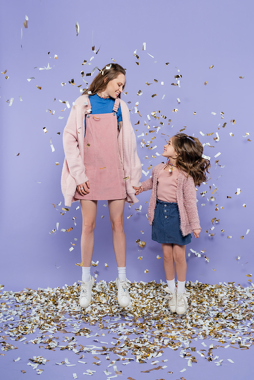 full length of happy mother and daughter holding hands and jumping near falling confetti on purple