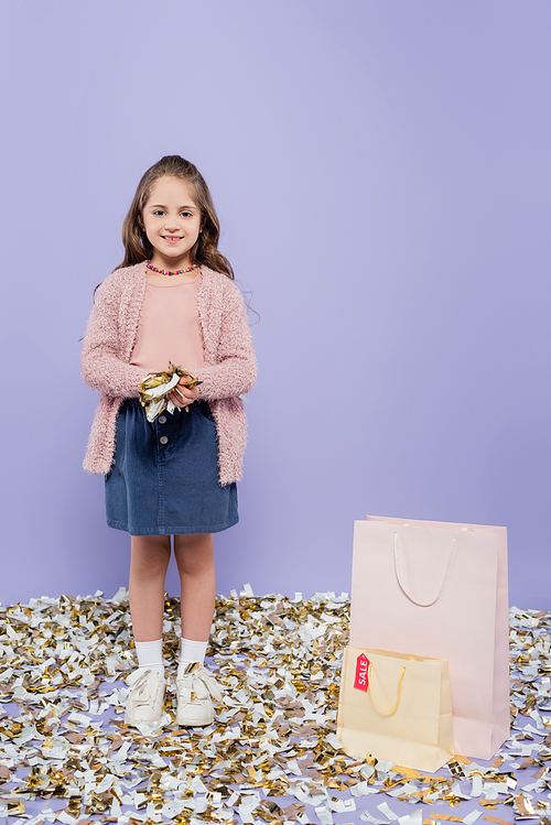 full length of happy girl holding confetti near shopping bags on purple