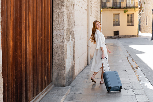 young cheerful woman in dress walking with suitcase on street in valencia