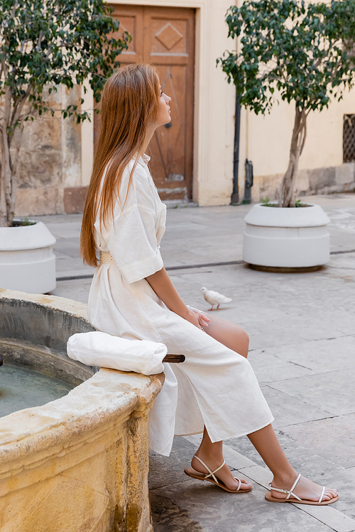 side view of redhead woman in white dress sitting near fountain on street of valencia