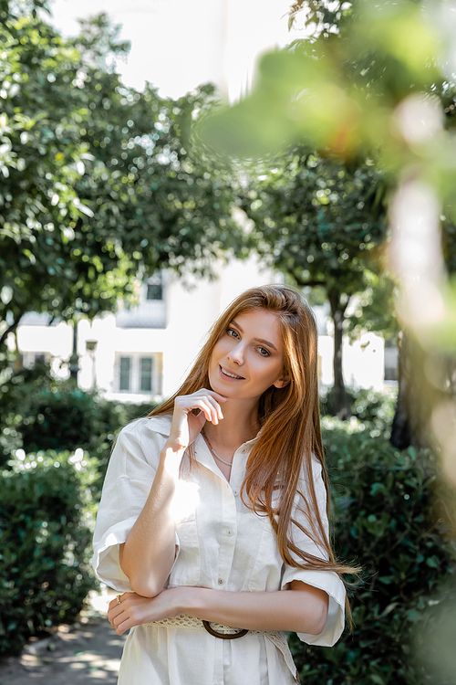 portrait of pleased and redhead woman in dress looking at camera in green park in valencia