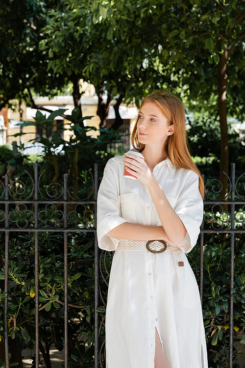 young redhead woman in white dress holding coffee to go near fence in european green park