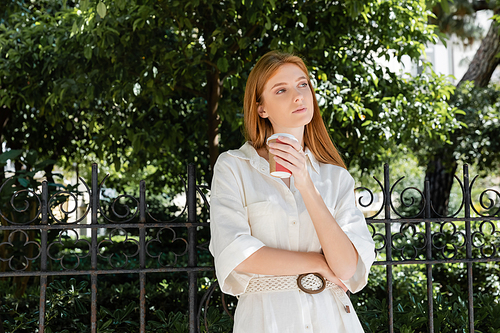 thoughtful redhead woman in white dress holding coffee to go near fence in european green park