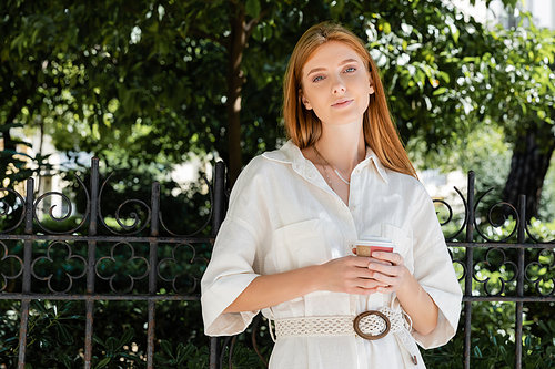 redhead woman in white dress holding coffee to go and looking at camera near fence in european green park