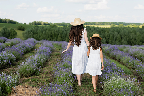 back view of mother and child with long hair and in white dresses walking in meadow