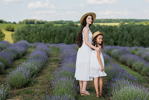 full length of smiling mom and daughter in white dresses looking at camera in blooming field