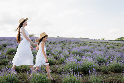 side view of mom and child in white dresses and straw hats holding hands and walking in field