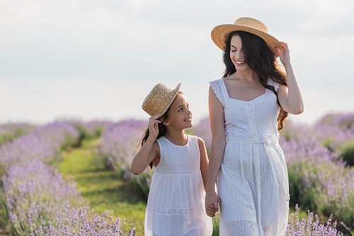 joyful mother and girl in straw hats holding hands and looking at each other in field