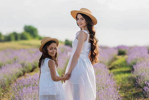 joyful girl and woman with long hair looking at camera and holding hands in meadow