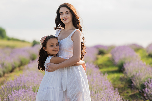 girl and mom in white dresses embracing and looking at camera in blurred field