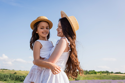 cheerful girl in straw hat looking at camera in hands of mom outdoors
