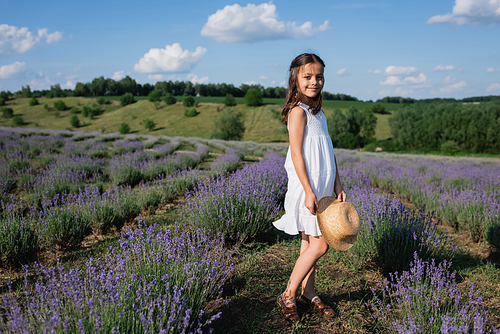 full length of smiling girl with straw hat standing in lavender field and looking at camera