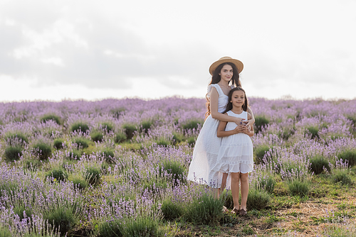 brunette woman in white dress and straw hat embracing daughter in flowering field